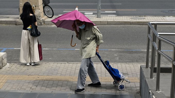 An elderly man walks along a sidewalk while holding an umbrella