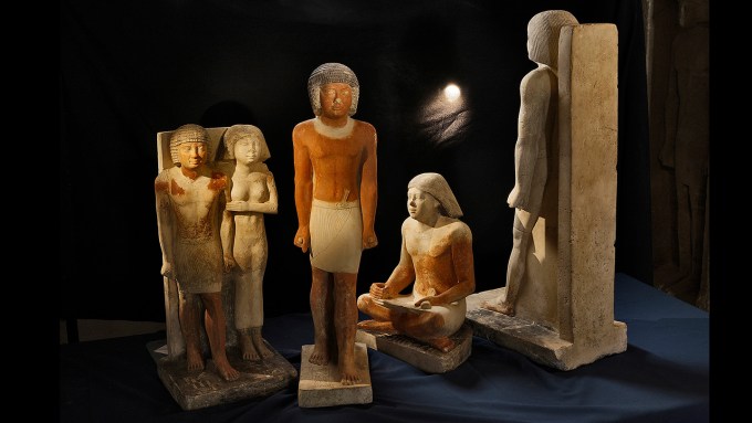 A series of stone statues on a dark backdrop depicting an ancient Egyptian scribe.