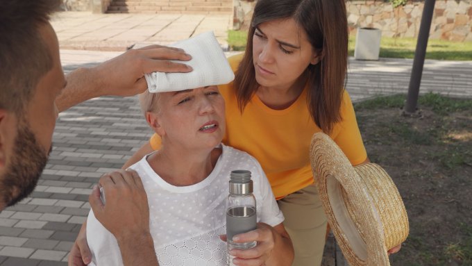 A man puts a white cloth on a woman's forehead. The woman is holding a water bottle and sitting in the shade as another woman standing behind her looks concerned