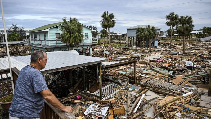 A man in a blue shirt stands on a deck and looks out over the vast landscape of destruction caused by Hurricane Helene.