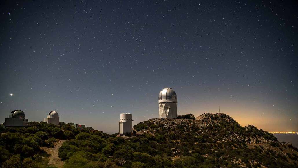 A line of four telescopes on a mountain ridge under a starry sky